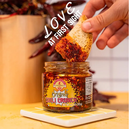 Jar of Original Chili Crunch with a hand dipping bread into it, set against a blurred indoor background.